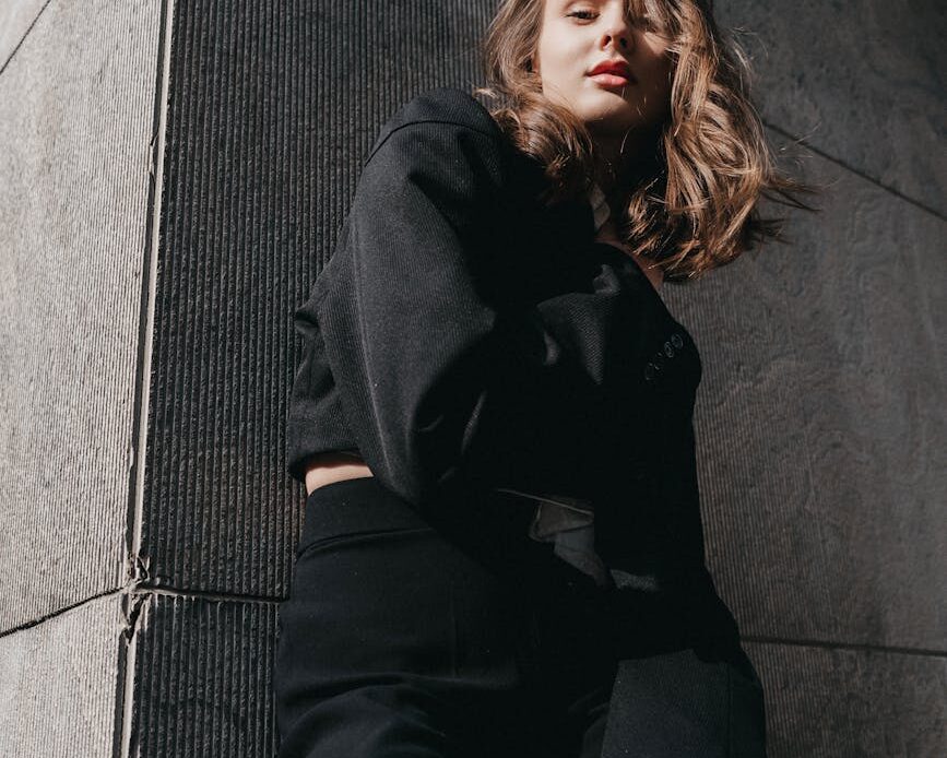 a woman in black pants and a black top leaning against a wall